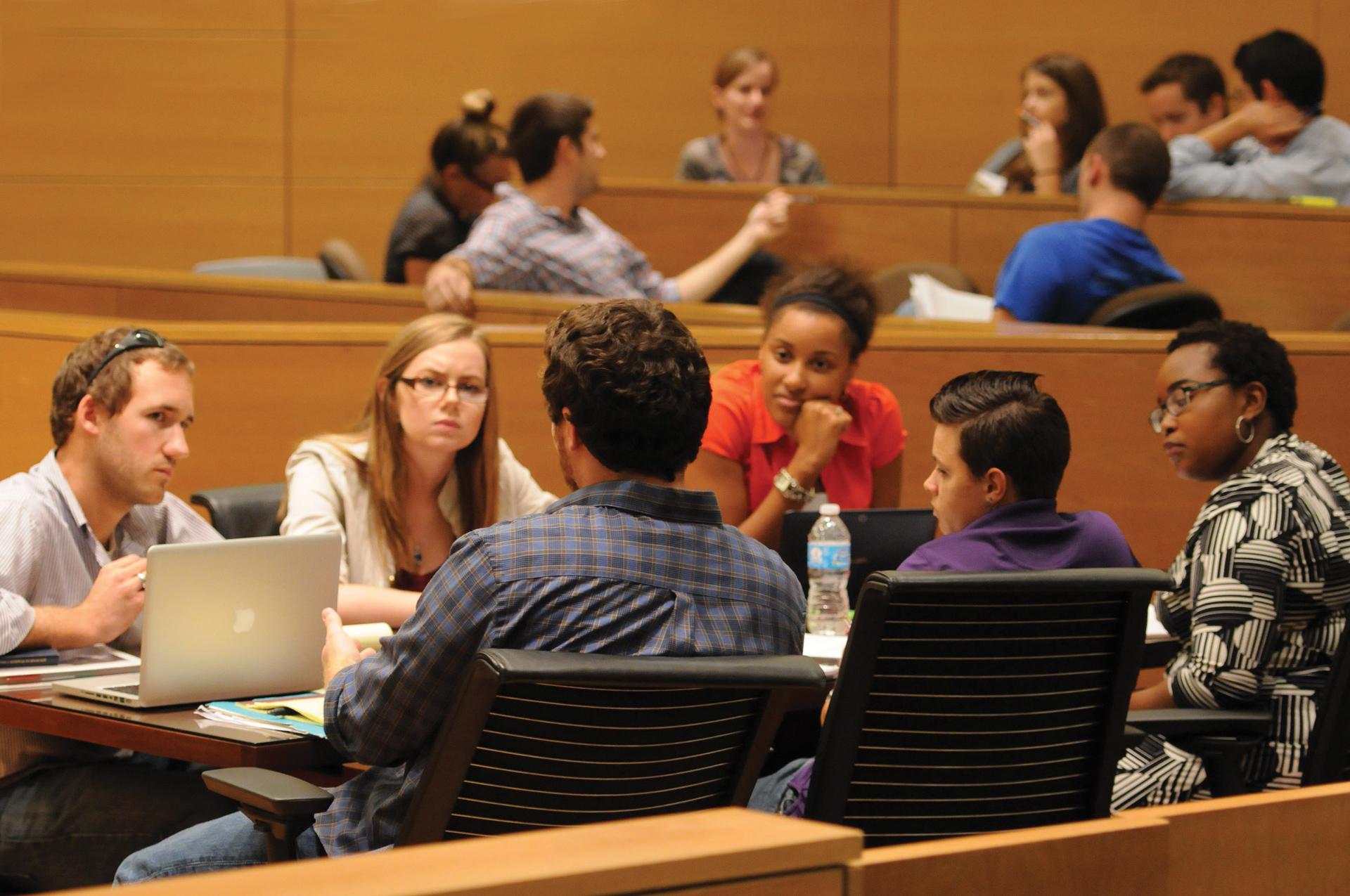 students sit at desks with computers while talking to each other