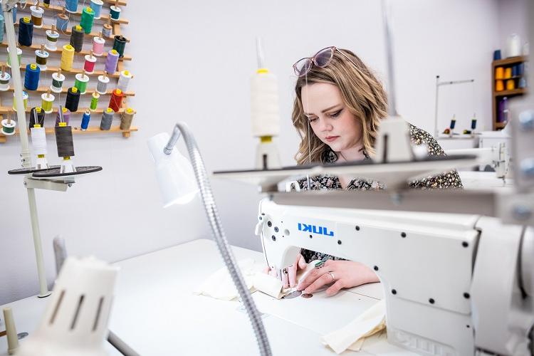 A student sits a sewing machine and works. On the wall beside her is a colorful assortment of thread.