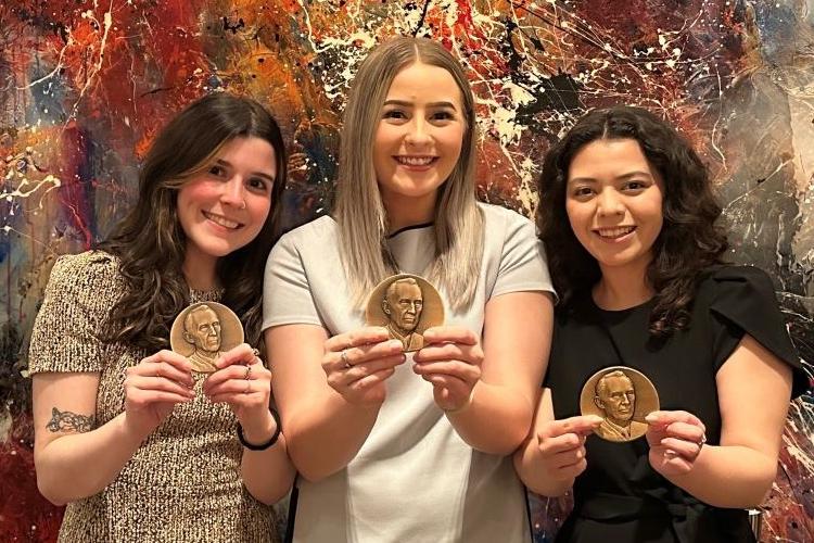 Martina Florido, Olivia Bess-Rhodes and Mattie Seley stand against a colorful wall, holding medals while smiling happily.